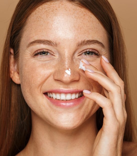 Beauty image of pretty shirtless woman smiling and applying face cream isolated over beige background